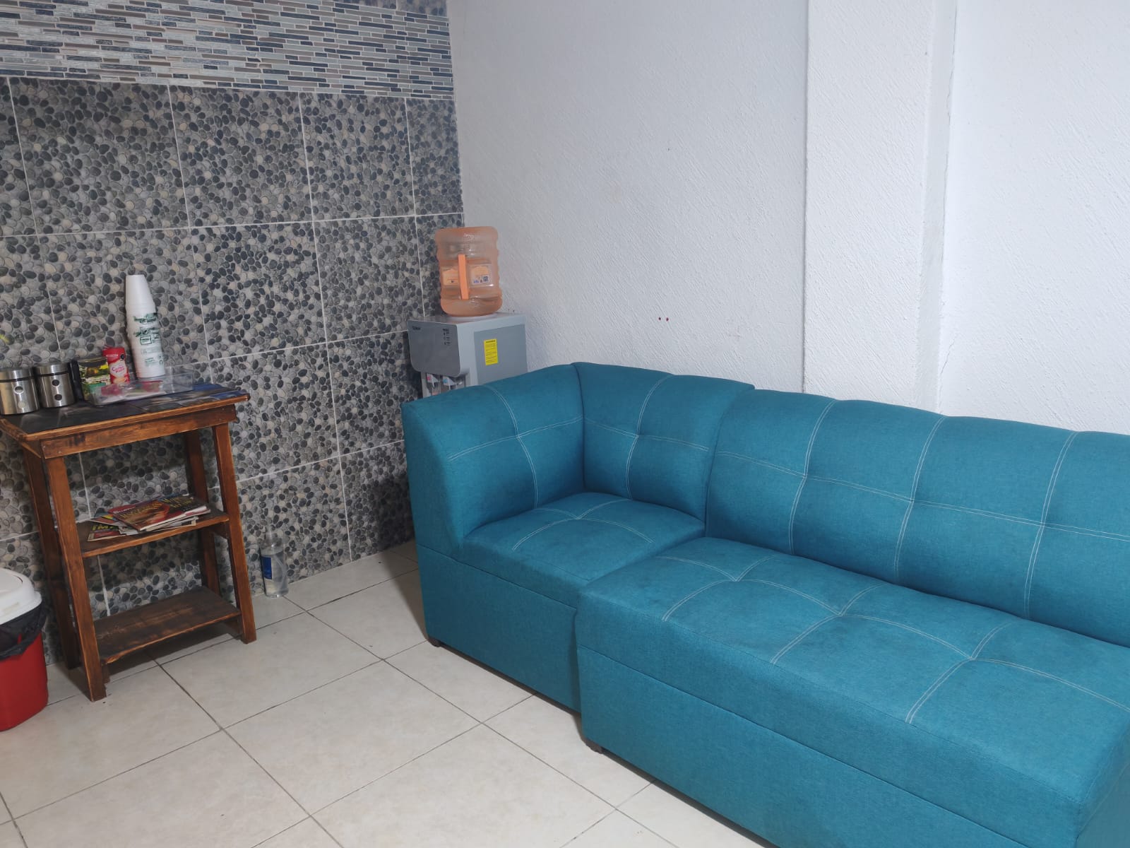 Sala de espera del consultorio de Calle Uvas, con sillón azul turquesa, muro de piedra y garrafón de agua